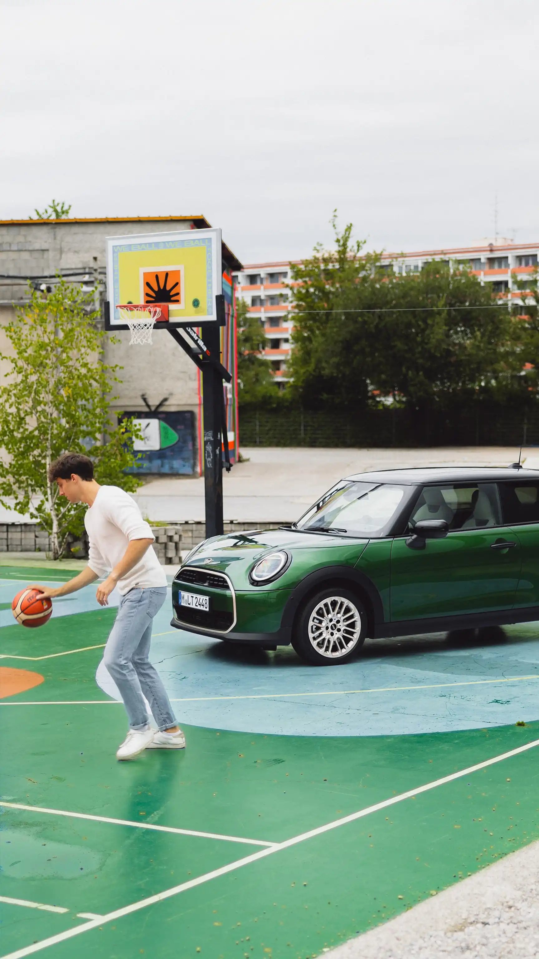 Человек ведёт баскетбольный мяч на открытой площадке рядом с MINI Cooper в цвете Racing Green IV.