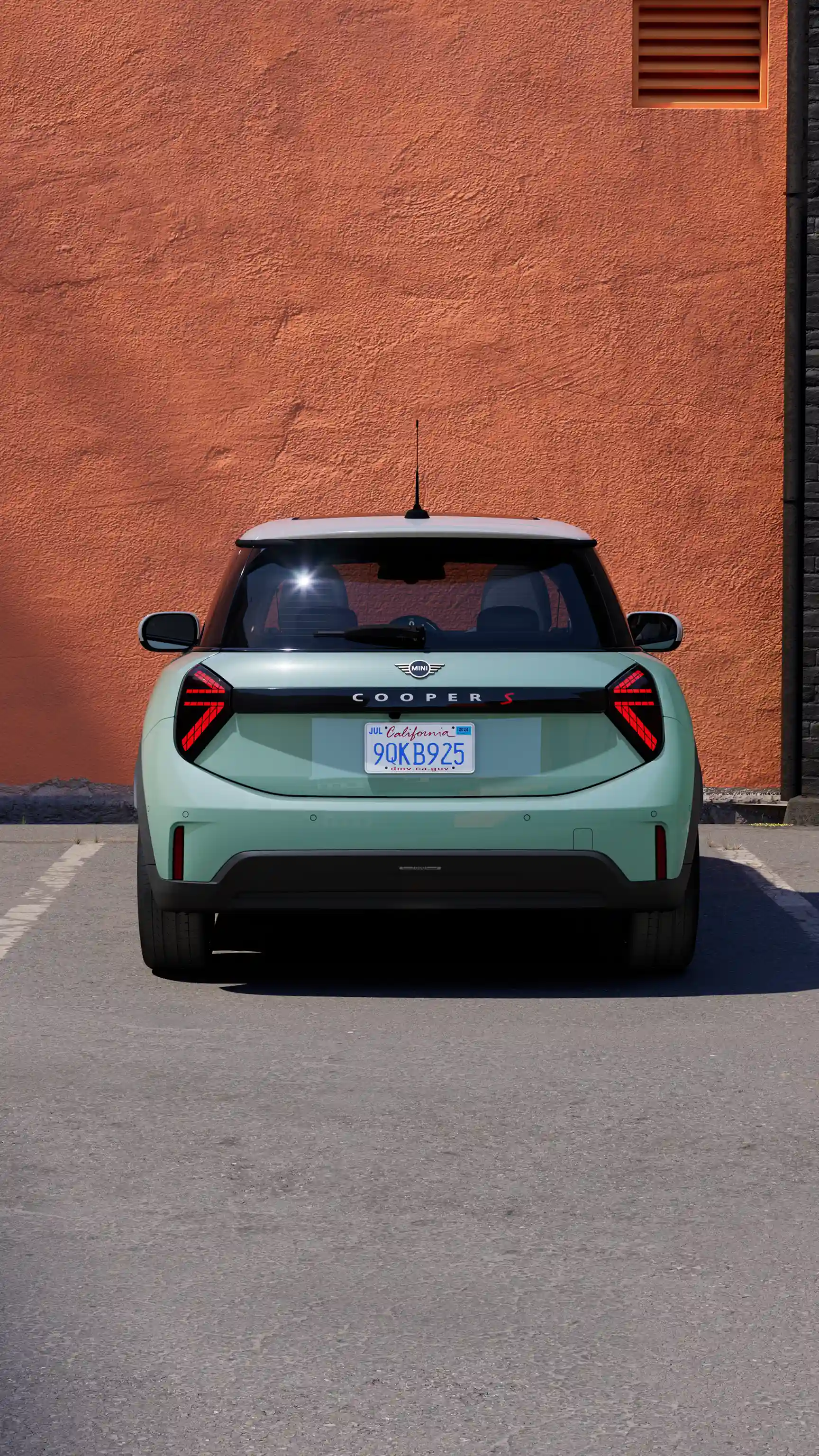 Вид сзади на MINI Cooper S в цвете Icy Sunshine Blue, припаркованный на размеченном парковочном месте у оранжевой стены.