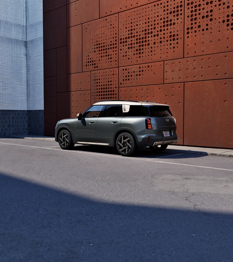 Вид сзади в три четверти на MINI Countryman в цвете Sage Green Metallic, припаркованный рядом с перфорированной стеной ржавого цвета.