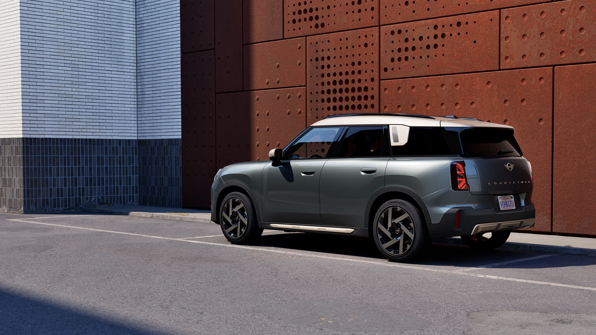 Вид сзади в три четверти на MINI Countryman в цвете Sage Green Metallic, припаркованный рядом с перфорированной стеной ржавого цвета.